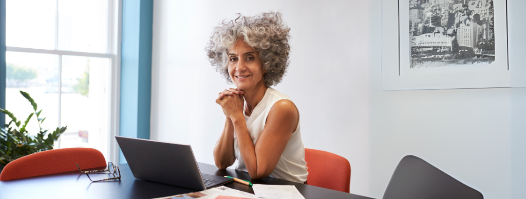 Smiling older woman with curly gray hair sitting at a desk with a laptop in a bright office.