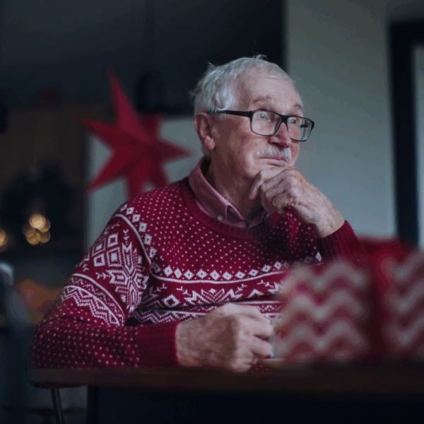 Older man in a festive red sweater sitting at a table, thoughtfully looking into the distance with soft holiday lights in the background